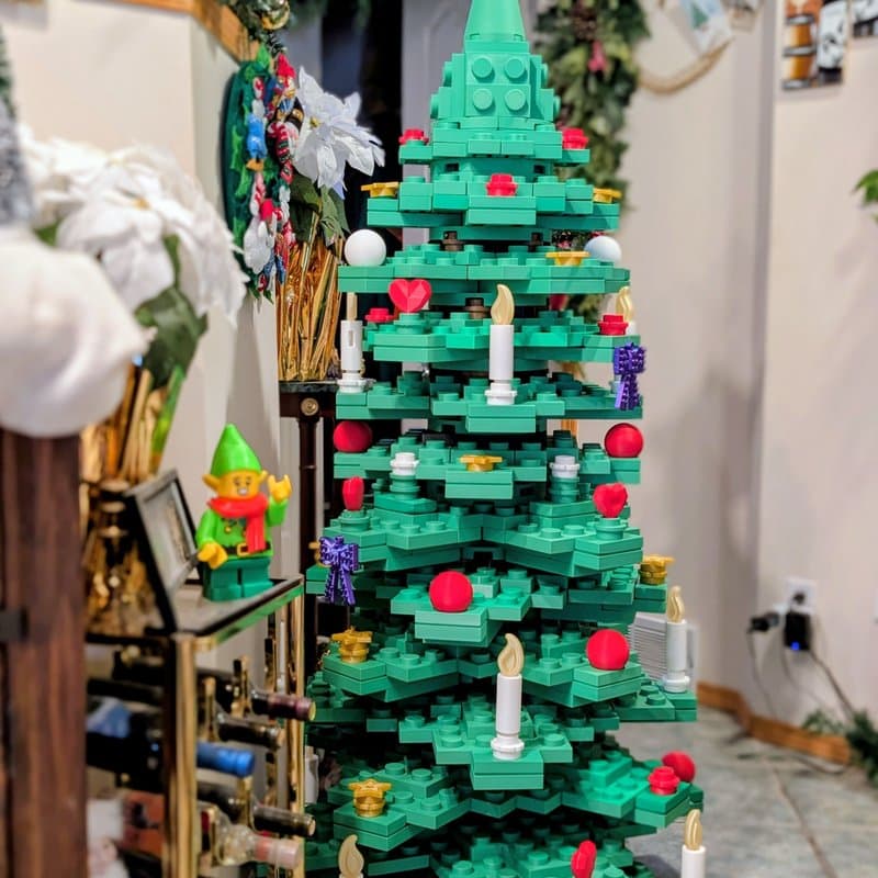 A colorful, Lego-style Christmas tree stands in a cozy indoor setting, adorned with red and white ornaments, candles, and a star on top. In the foreground, a small Lego elf figure with a green hat and red scarf is positioned next to a wine rack filled with bottles.